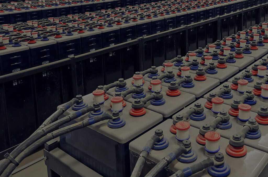 A large array of industrial batteries is arranged in rows, connected by numerous cables and wires. The batteries, featuring red and blue components on top with black casings below, are neatly organized behind a perforated plate in what appears to be a storage or power facility.