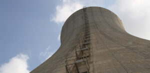 A close-up view of a large concrete cooling tower against a backdrop of a partly cloudy sky, with a metal ladder and platform attached to the side stretching upwards towards the top of the structure.