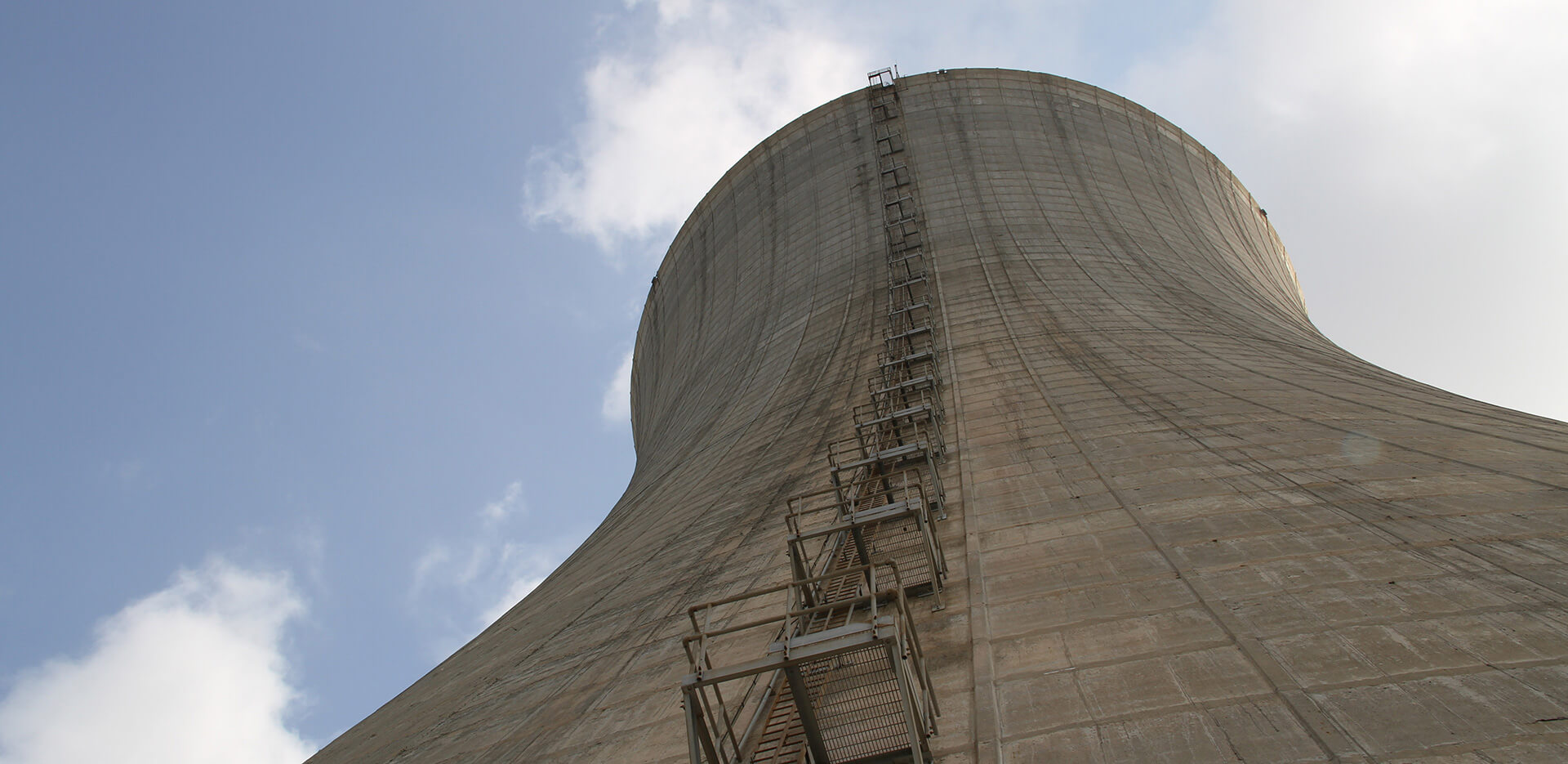 A close-up view of a large concrete cooling tower against a backdrop of a partly cloudy sky, with a metal ladder and platform attached to the side stretching upwards towards the top of the structure.