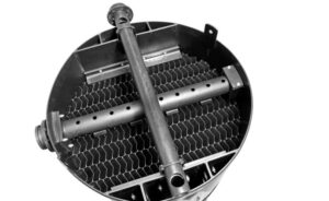 Close-up of a cylindrical industrial component resembling a sedimentation tank with a grid-like structure inside. Two horizontal pipes with multiple holes run through the center of the cylinder, and there are several connectors and brackets attached to the edges.
