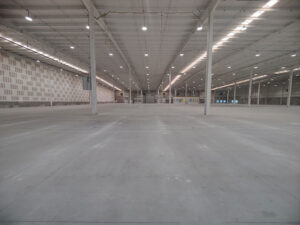 A vast empty warehouse with a high ceiling, bright overhead lighting, and expansive concrete flooring. The interior features exposed beams and support columns.