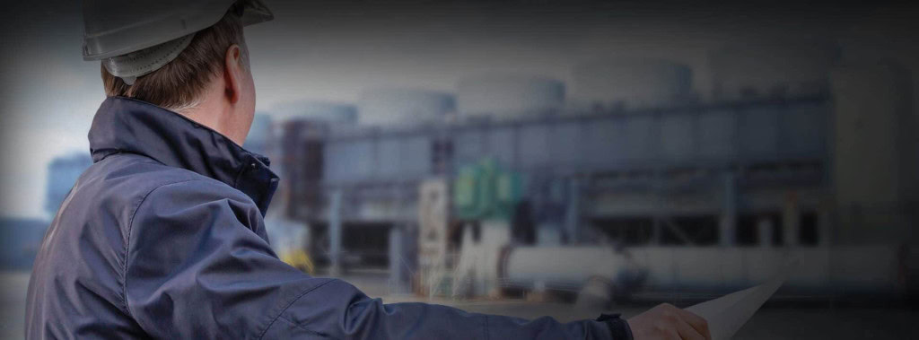 Person in a protective helmet and jacket, viewed from behind, looking at an industrial facility with large cylindrical structures and various equipment in the background.