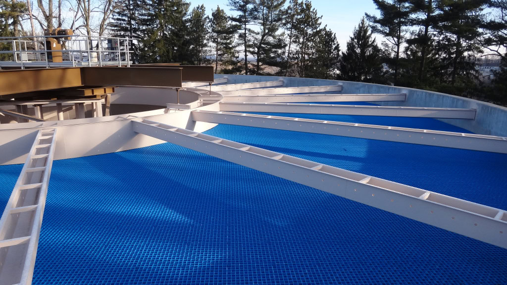 Estructura industrial circular con vigas en ángulo que sostienen una rejilla azul en el suelo, parte de un sistema de tratamiento de aguas o de filtración de aguas residuales. De fondo, altos árboles bajo un cielo despejado.