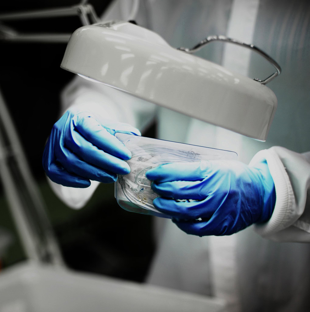 a person wearing blue gloves and a white protective lab coat, working under a bright lamp, holding a clear plastic container