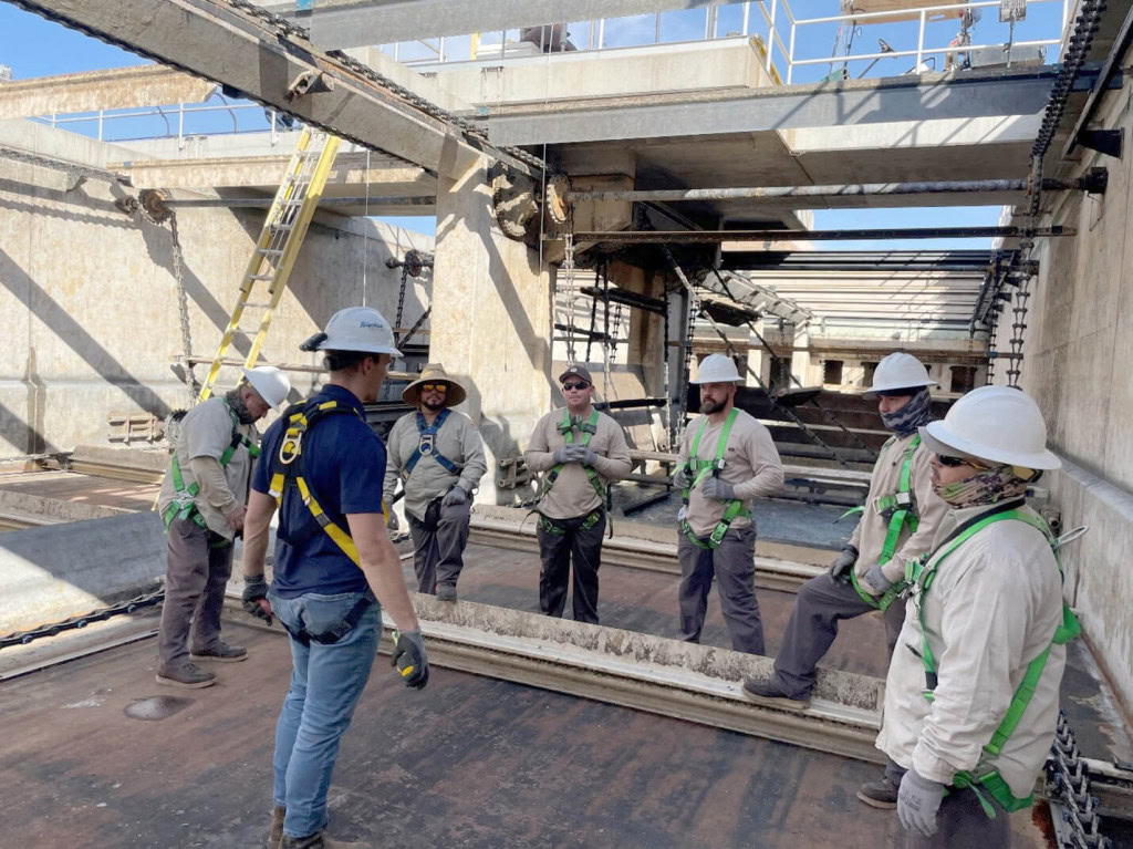 Un grupo de trabajadores de la construcción con equipo de seguridad, incluidos cascos y arneses, de pie en una obra. Un trabajador mira al grupo, aparentando dar instrucciones.
