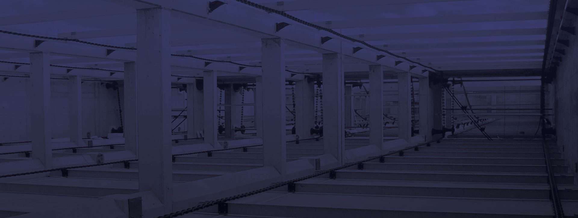 Un escenario interior oscuro e industrial con grandes vigas metálicas y cadenas suspendidas del techo. La sala está vacía en su mayor parte, con filas visibles de plataformas y una estructura en forma de rejilla encima.