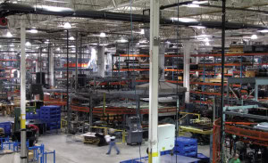 A large industrial warehouse with shelves stocked with various materials and equipment. The manufacturing space features high ceilings with exposed beams and bright lighting.