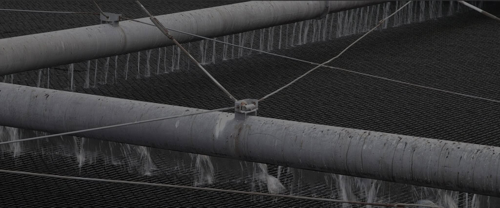 Close-up of large industrial pipes with water cascading through a metal mesh structure. The pipes, carrying wastewater, are supported by a network of cables, creating a complex yet organized arrangement.