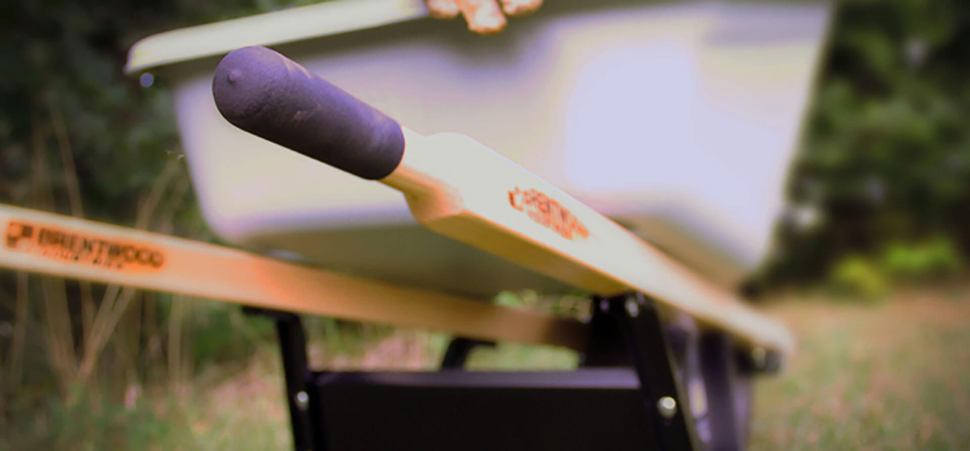 A close-up shot of the handle and upper body of a Brentwood wheelbarrow on a grassy surface. The image focuses on the handle's wooden grip and a portion of the metal tub, with a blurred background of greenery.