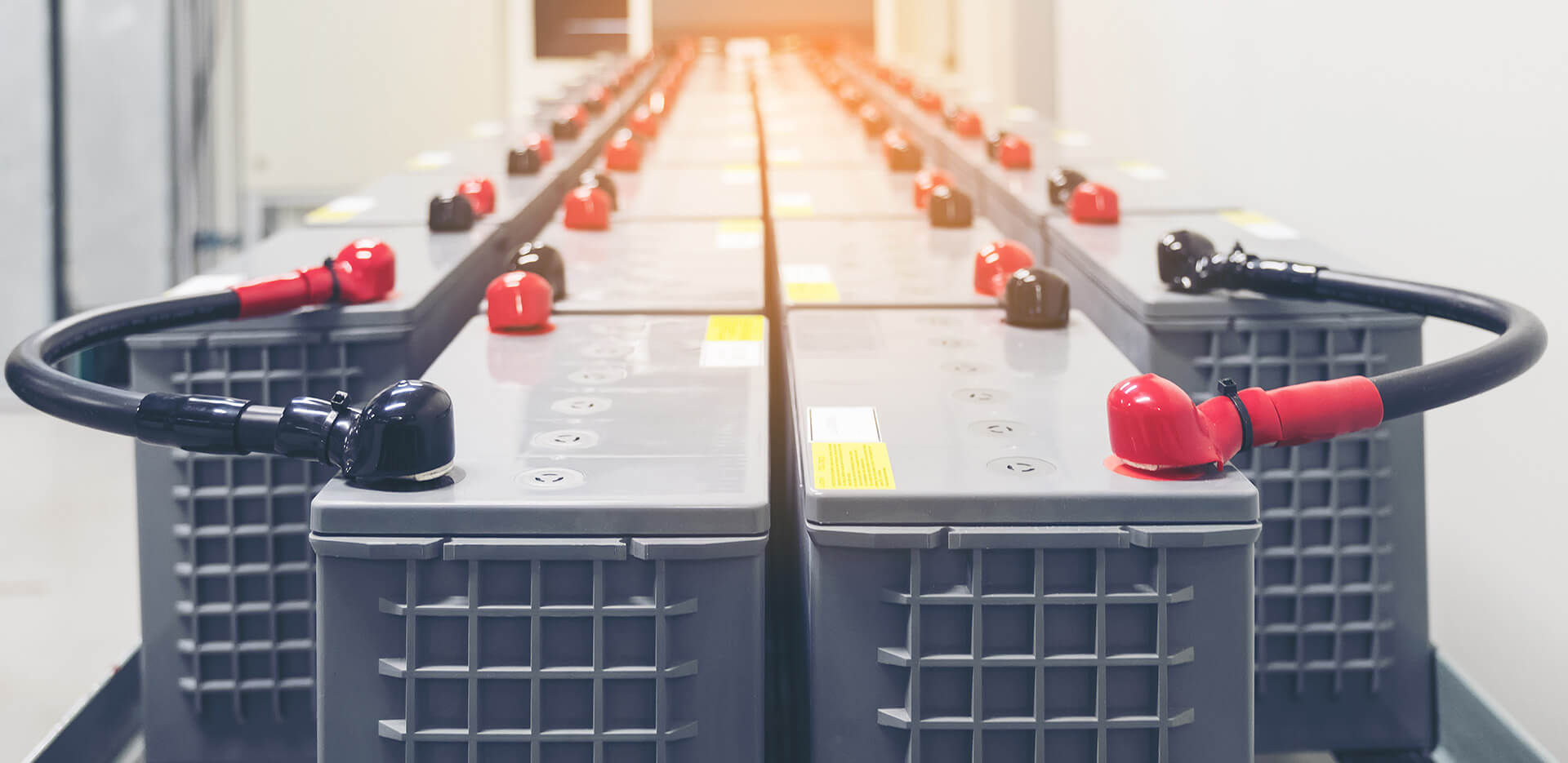 A row of industrial batteries arranged symmetrically in a brightly lit room. Each battery is fitted with red and black terminals, with cables attached.