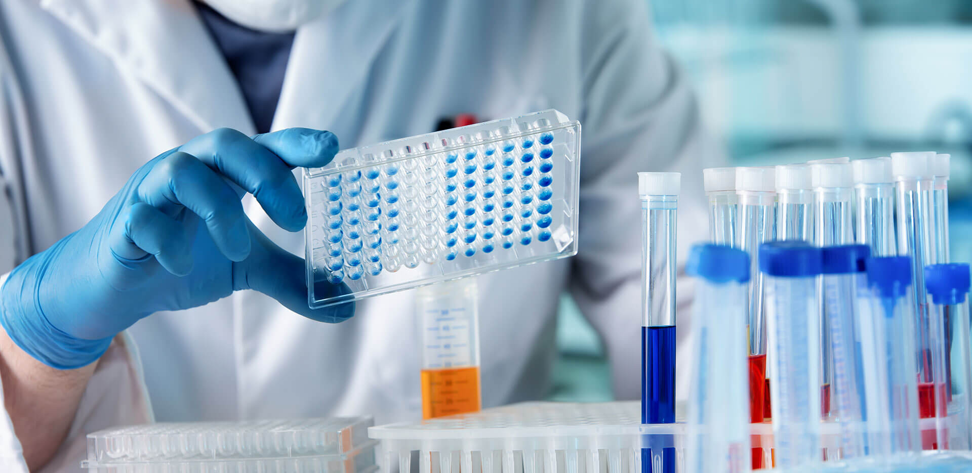 A scientist wearing a lab coat and blue gloves holds a multi-well plate filled with samples. Various test tubes containing colorful liquids are organized on the lab bench in the foreground.