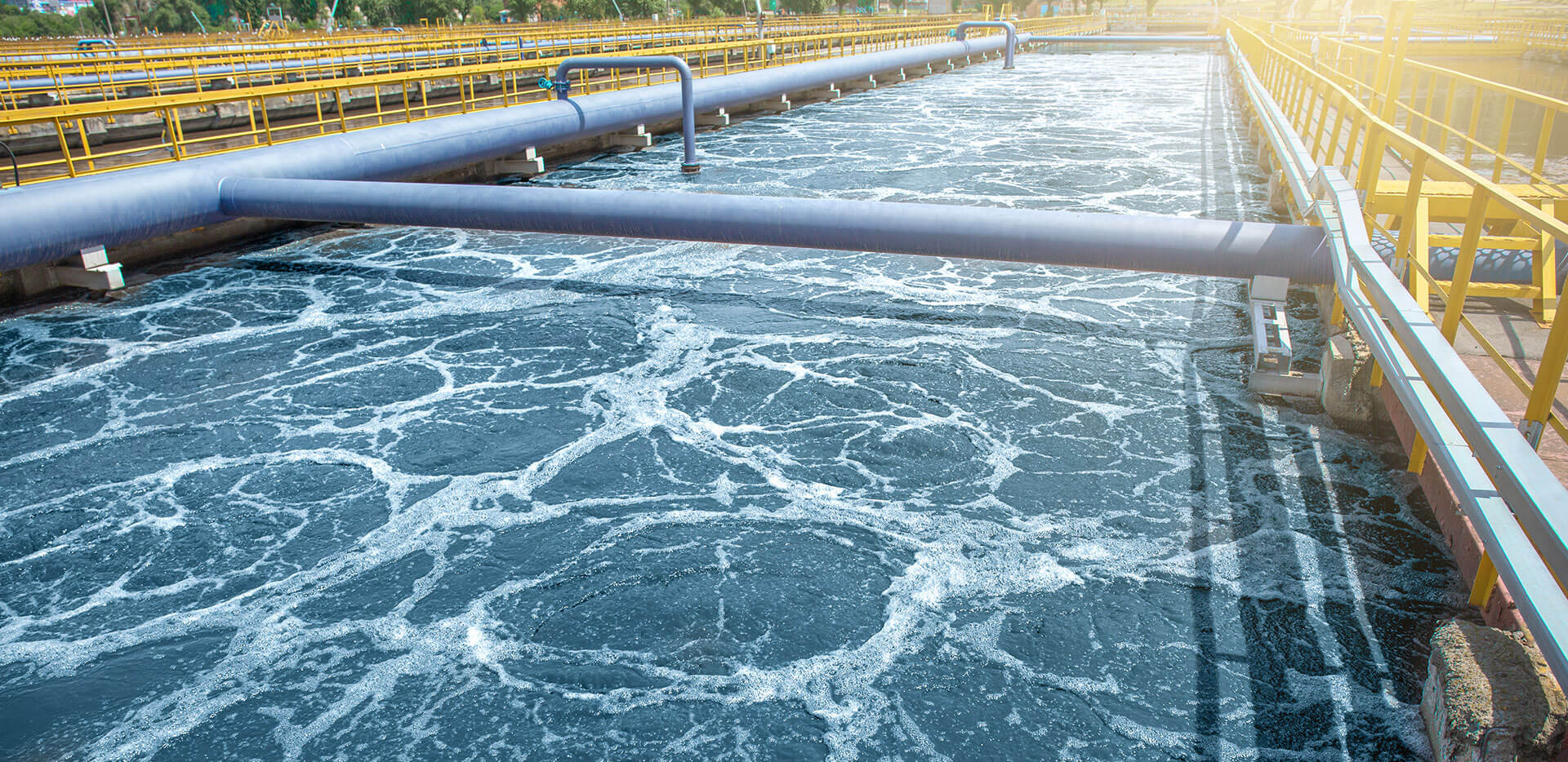 A large industrial water treatment facility with aerated water in a long, rectangular tank. Multiple pipes and yellow railings run alongside the water.