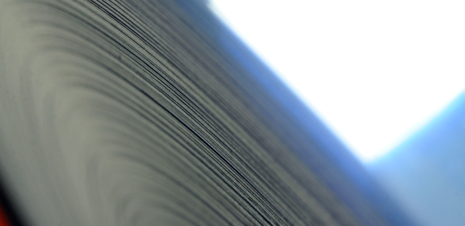 Close-up of the edge of a large stack of paper or a roll of paper, with the tightly packed sheets creating a series of curved lines. The lighting is soft, evoking a warm home feel, with a blue tone on the left and a brighter, whiter light on the right.