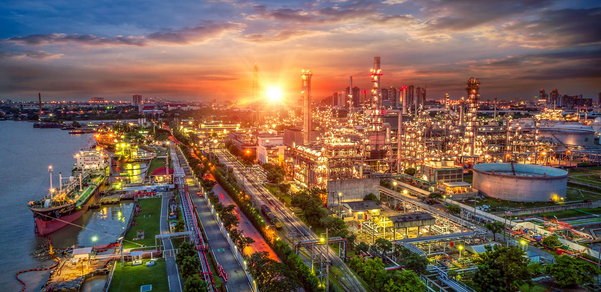 A sprawling industrial complex with numerous brightly lit structures and tanks is seen next to a river, under a sunset sky.